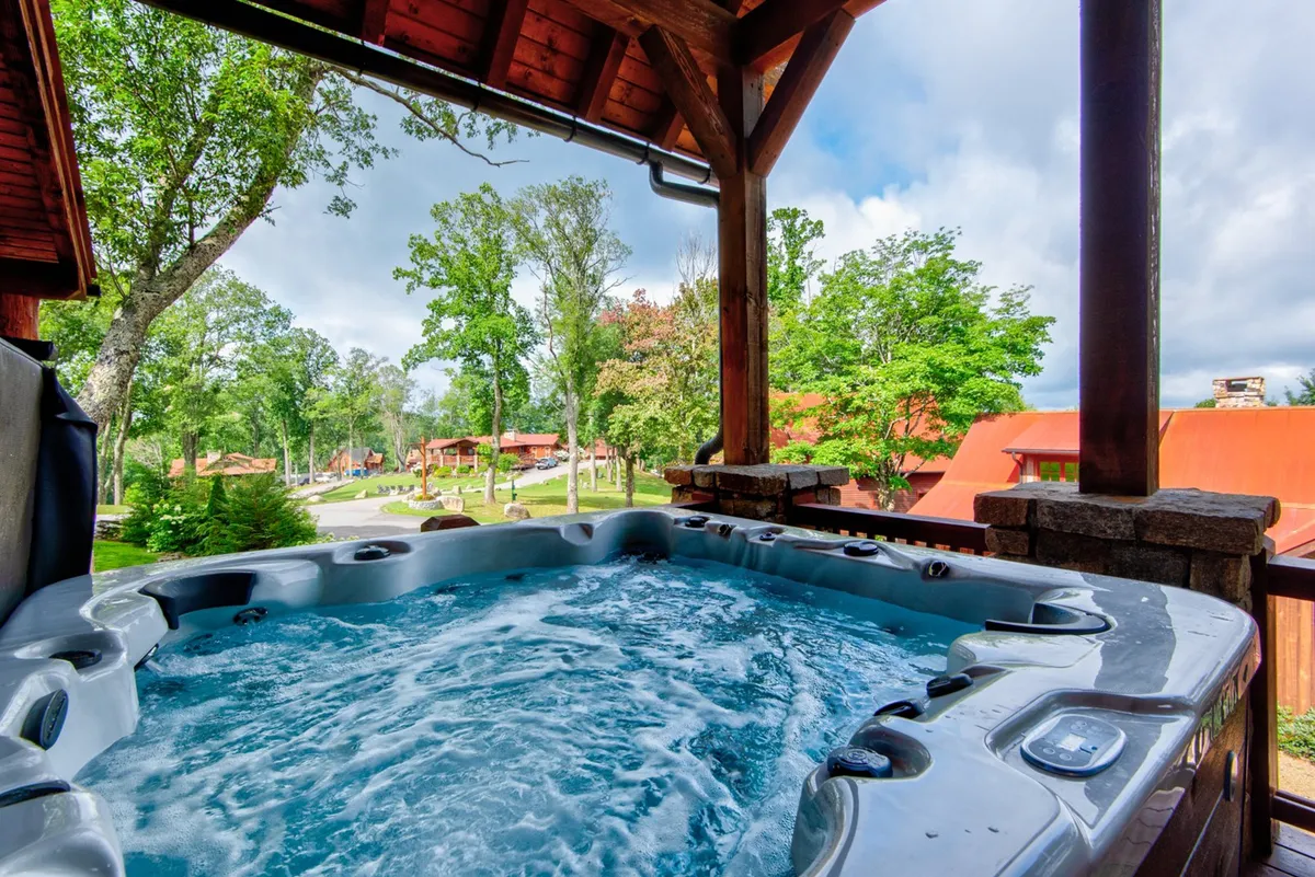 The image features a bubbling hot tub set on a wooden deck surrounded by tall trees, with a partial view of nearby cabins. The scene is tranquil, suggesting a private, nature-immersed space perfect for relaxation and stargazing.