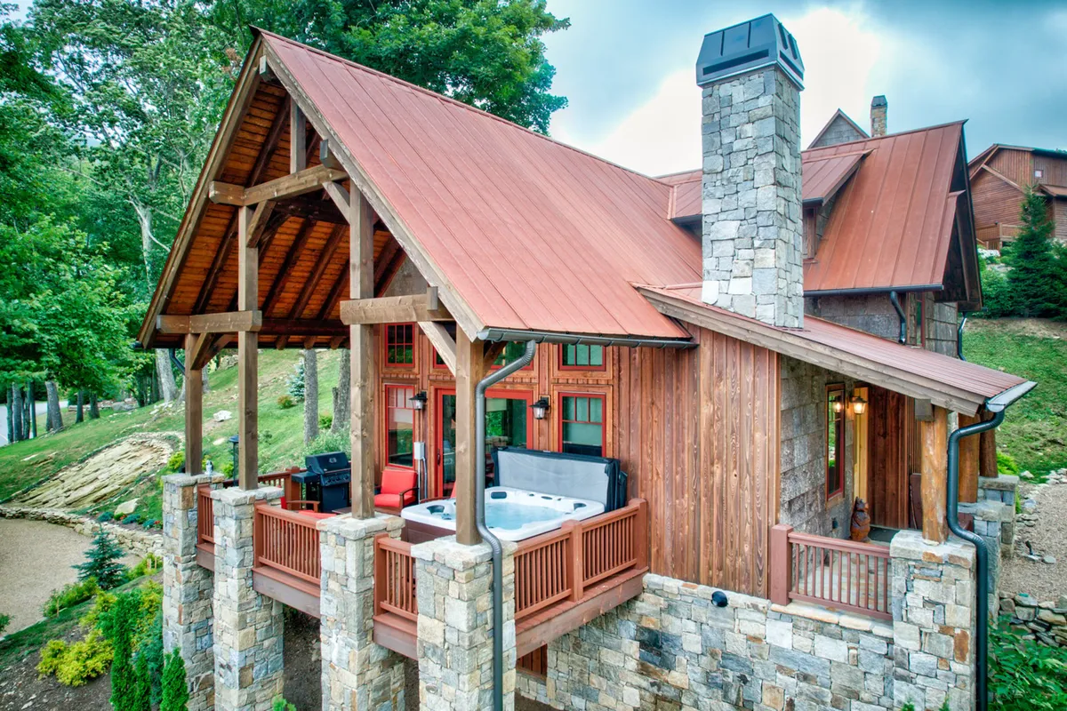 This image captures a wide view of the stone chimney, wooden beams, a spacious deck with an exterior seating area, and a hot tub. The composition highlights the outdoor living space and its seamless integration with nature, ideal for gatherings and relaxation.