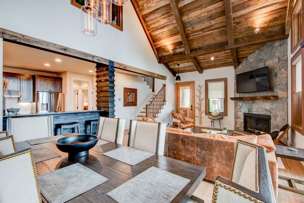 A warm, inviting interior with a large dining table set near a stone fireplace and under rustic wooden beams. The space also features a visible staircase and elements of the bar area, capturing the seamless flow between living and dining zones. The ambiance suggests an ideal spot for sharing meals, celebrating special occasions, or enjoying quiet, fireside moments.