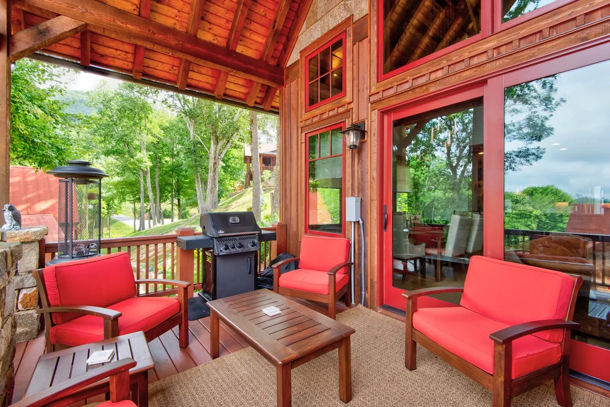 An outdoor setting with red armchairs, a wooden table, and a grill, viewed against a backdrop of large windows and scenic greenery. The area feels intimate and is clearly designed for socializing, dining, and celebrating in a relaxed, natural environment.