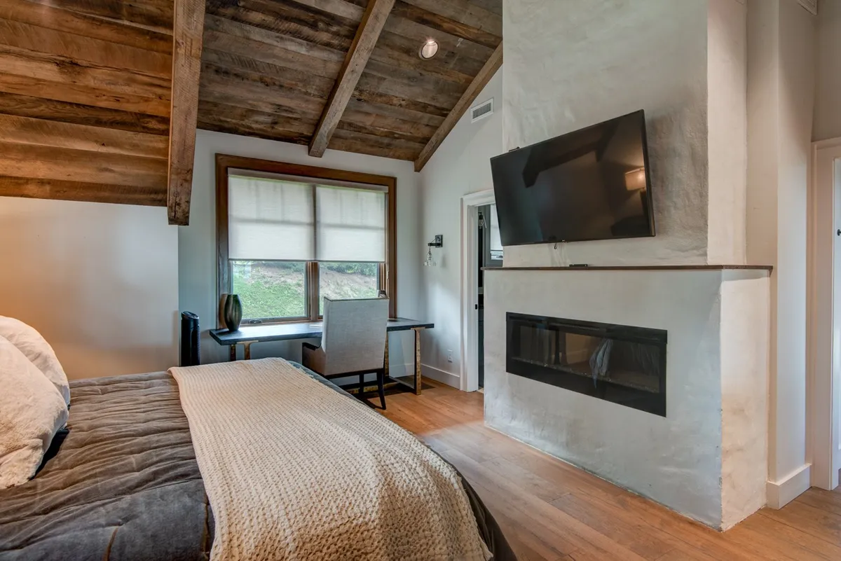 A serene king bedroom with a cozy bed, a window that lets in natural light, a small desk, a fireplace, and a television. The space is arranged for relaxation and quiet evenings, with clean lines and calming colors that foster a peaceful environment.