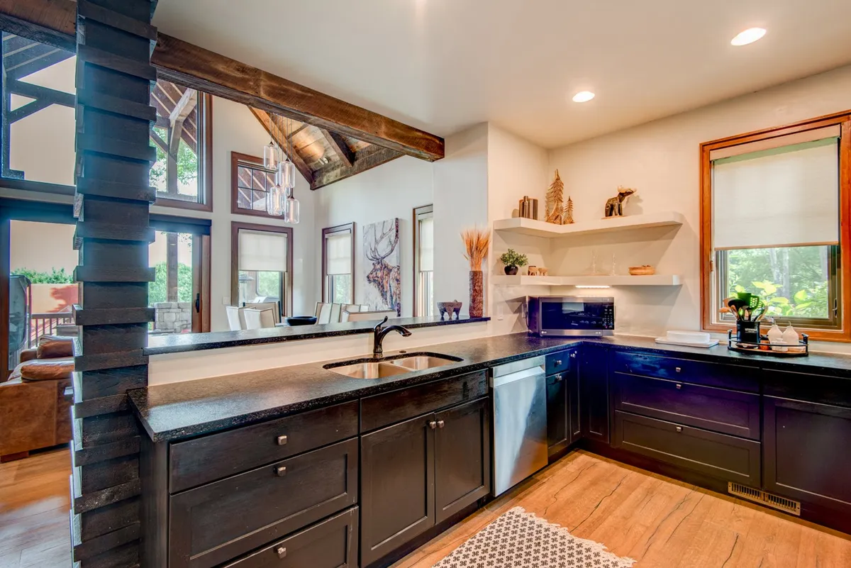 A bright, welcoming kitchen space with dark cabinets, an expansive stainless steel sink, open shelving stocked with kitchen essentials, and large windows that flood the room with natural light. Wooden beams and a mountain-modern aesthetic connect the kitchen to its scenic locale.