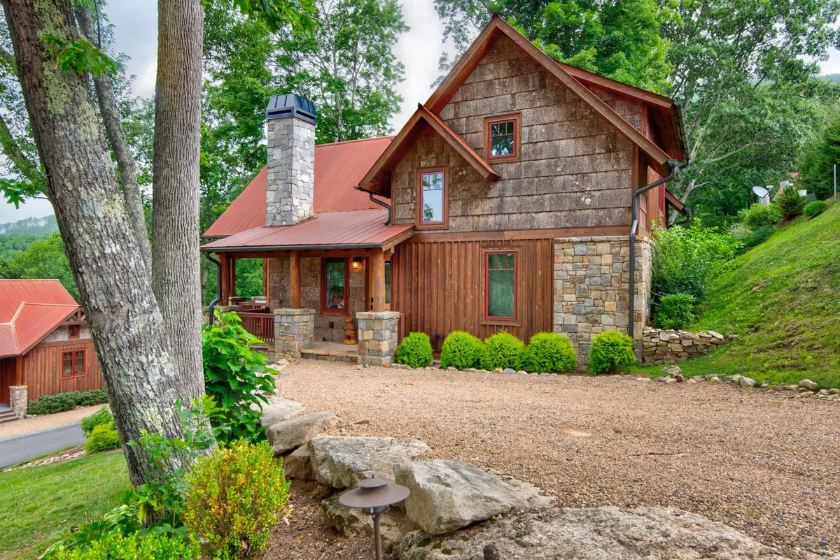 The image showcases a charming wooden cottage with a stone chimney nestled among lush greenery. A gravel pathway leads to the inviting front porch, which is ideal for enjoying sunset views or stargazing in the evenings. The cozy ambiance is enhanced by seasonal decorations, and the overall scene radiates warmth and tranquility, perfect for outdoor gatherings or quiet relaxation.