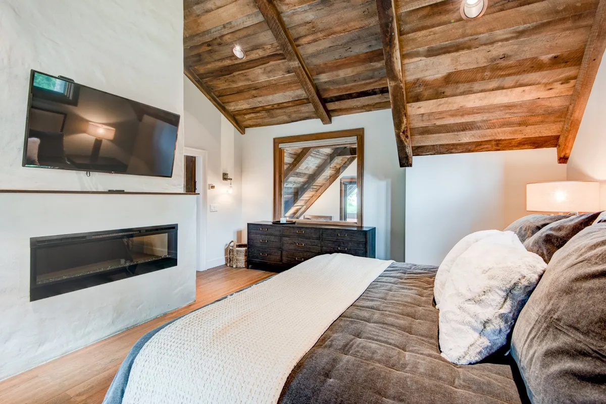 A spacious, inviting king bedroom featuring plush bedding, exposed wooden beams, a large mirror, a wall-mounted television, and a stone fireplace. The room exudes comfort and warmth, with soft lighting and thoughtful touches that create a restful, romantic ambiance. The fireplace and TV echo the modern comforts mentioned, while the high-quality linens and cozy decor provide a sense of luxury and privacy.