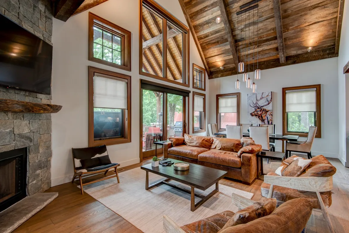 A welcoming open-plan living and dining area bathed in natural light, featuring a rustic stone fireplace as the focal point. The cozy leather sofa sits across from the hearth, while the modern dining area, complete with stylish seating and artwork, extends into view. Large windows frame the space, allowing sunlight to highlight the warmth of the oak floors and exposed barn-wood ceiling beams.