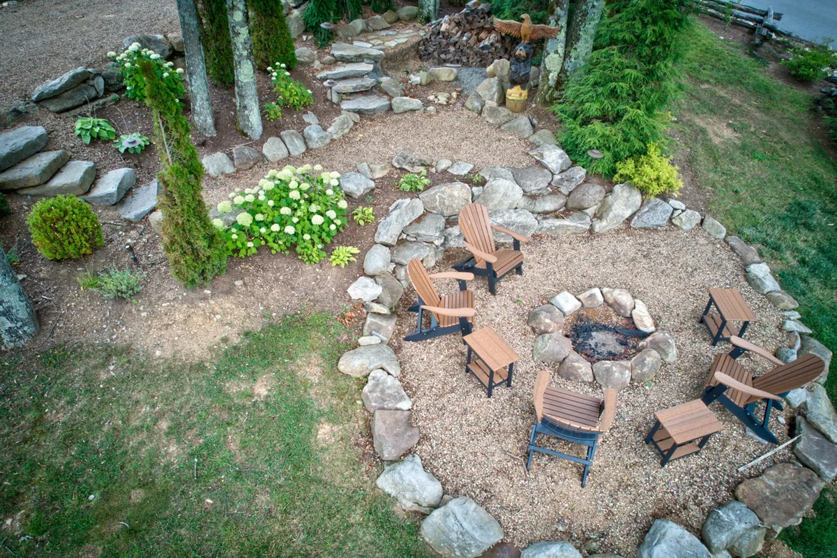 A cozy stone fire pit is encircled by wooden chairs and surrounded by flower beds and a stone pathway. The setting is warm and inviting, perfect for evening gatherings, storytelling, or roasting marshmallows under the stars.