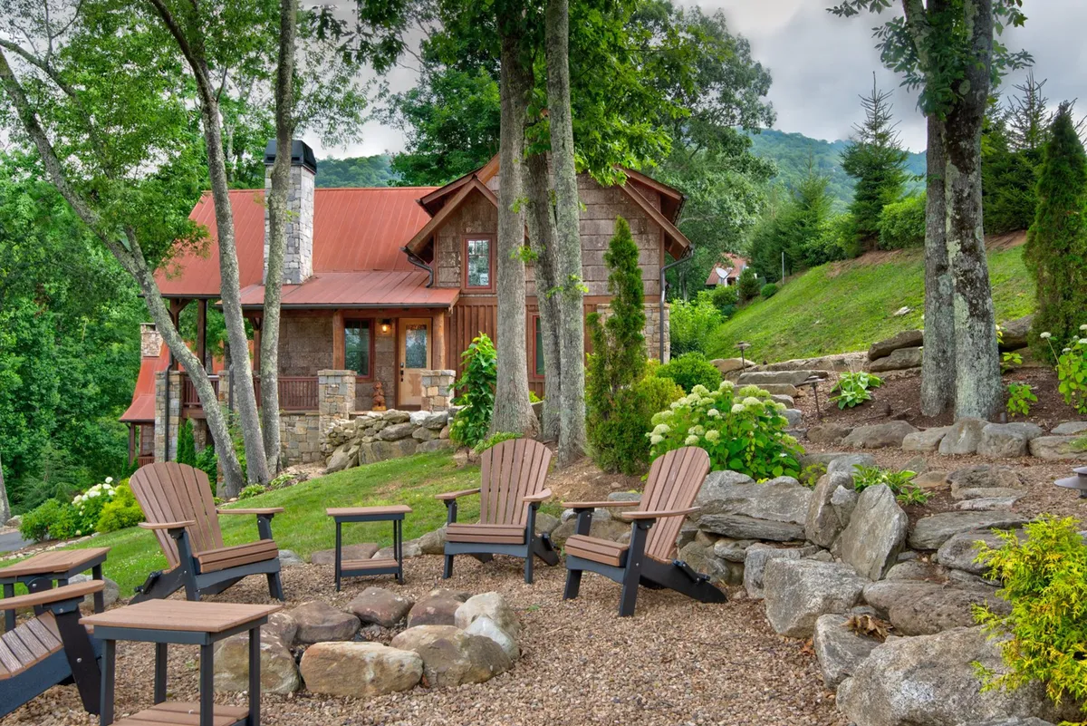 A cozy fire pit setup with Adirondack chairs encircling the fire, a landscaped garden, and a charming house with a red roof in the background. The atmosphere is inviting and intimate, ideal for evening gatherings and quiet moments under the stars. The stone pathway and lush garden enhance the sense of tranquility and retreat.