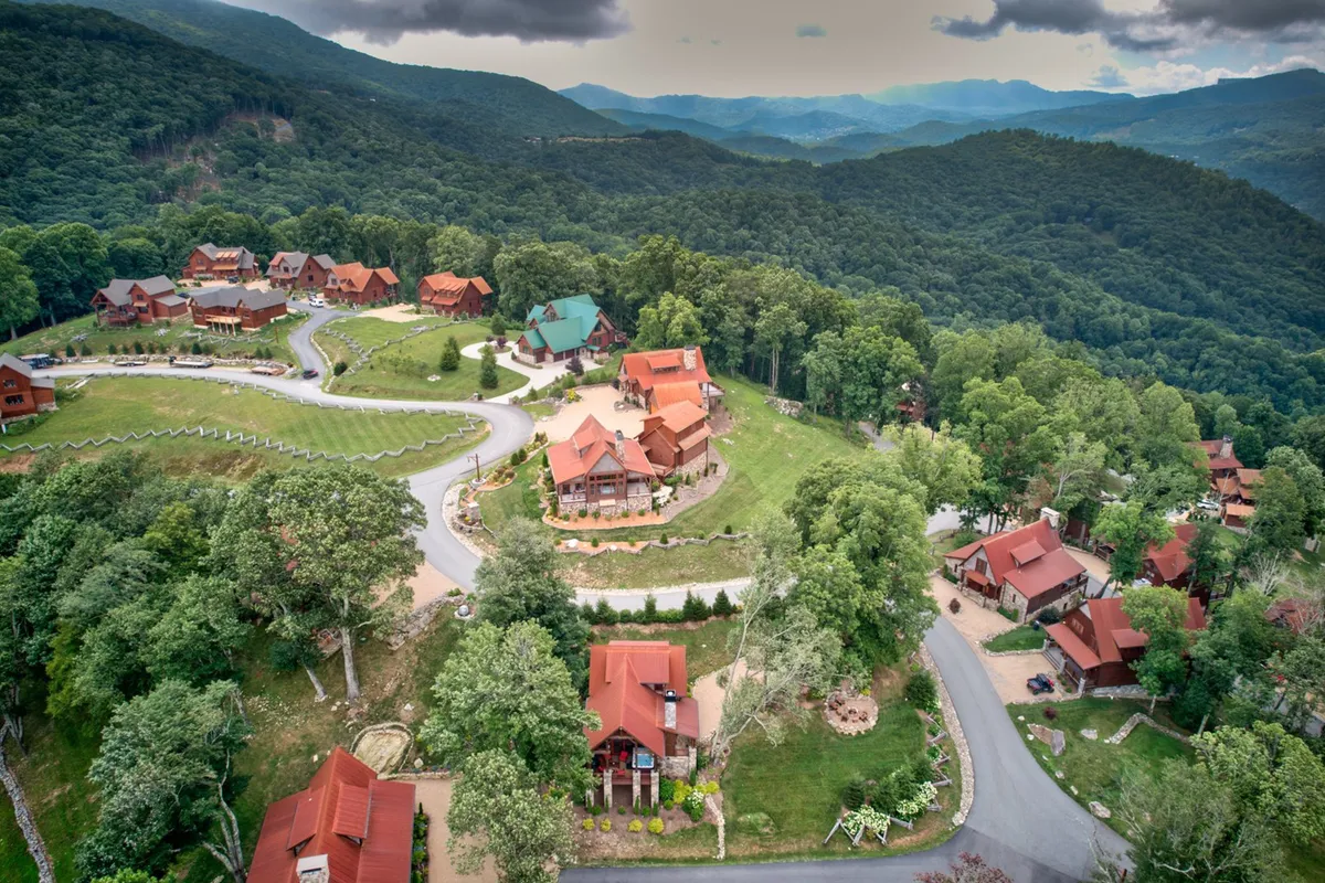 This exterior shot features charming log cabins nestled among rolling green hills and lush trees, with winding roads inviting exploration. The landscape suggests open areas ideal for nature walks, family hikes, and community-based outdoor activities. The serene sunset hints at family evenings spent together outdoors, possibly enjoying events or storytelling.