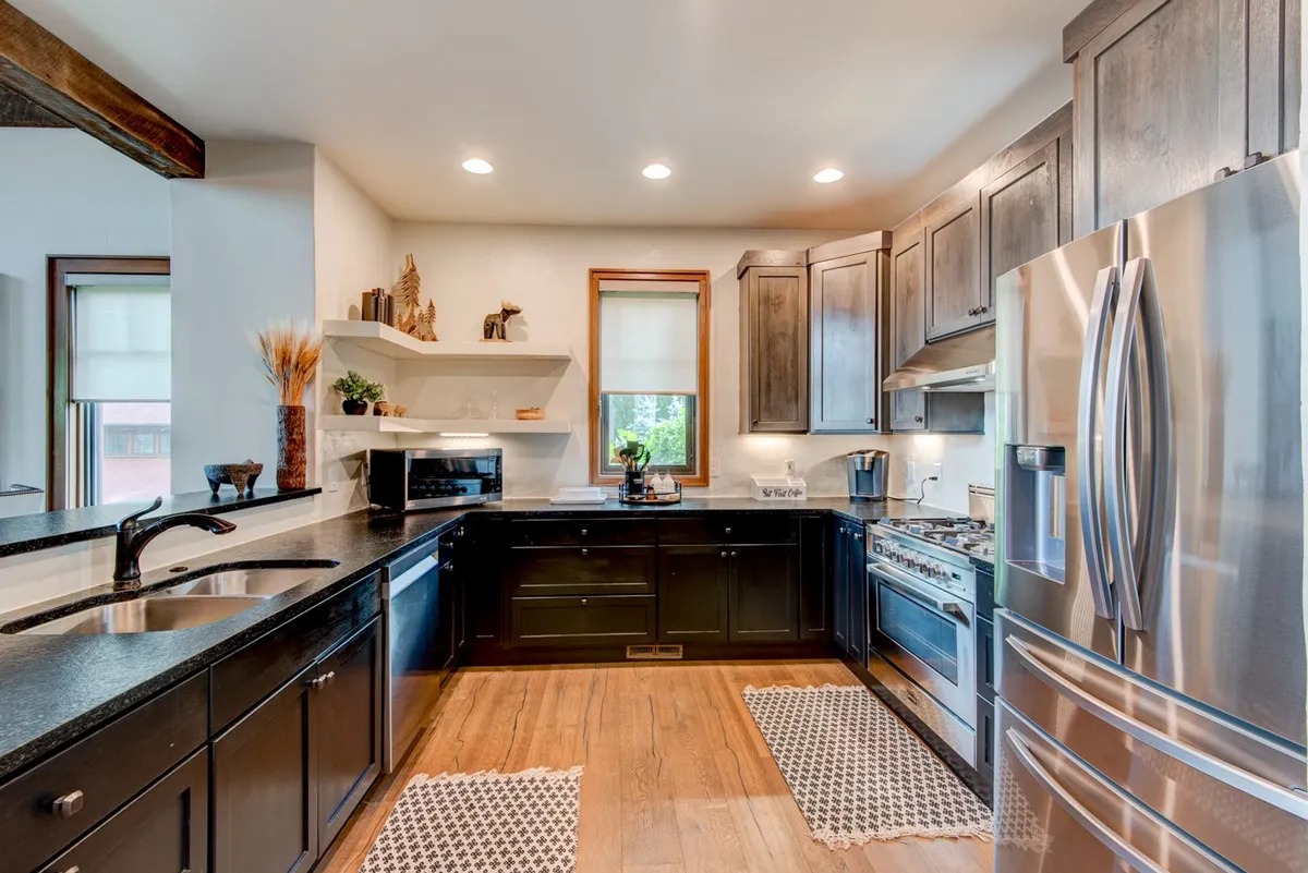A cozy, functional kitchen featuring striking black cabinetry, stainless steel appliances, a warm wooden countertop, and decorative shelves with kitchenware. The styling hints at a blend of rustic charm and modern convenience, with everything needed for meal prep and entertaining within reach.