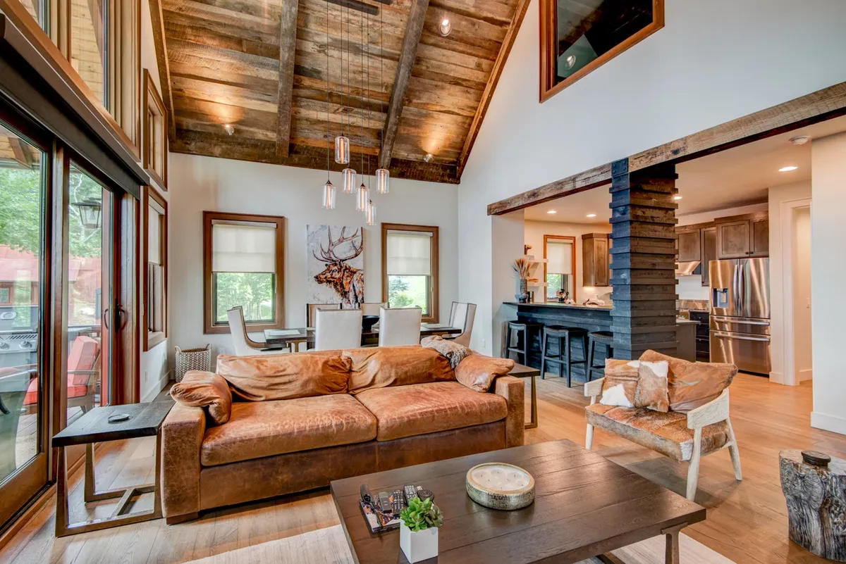 An elegant, open-concept interior highlighted by a leather sofa, exposed wooden beams on the ceiling, and a spacious dining area adjacent to a kitchen island. The cozy mood is enhanced by warm accent lighting, and the entire setting invites relaxation, socializing, and group activities.