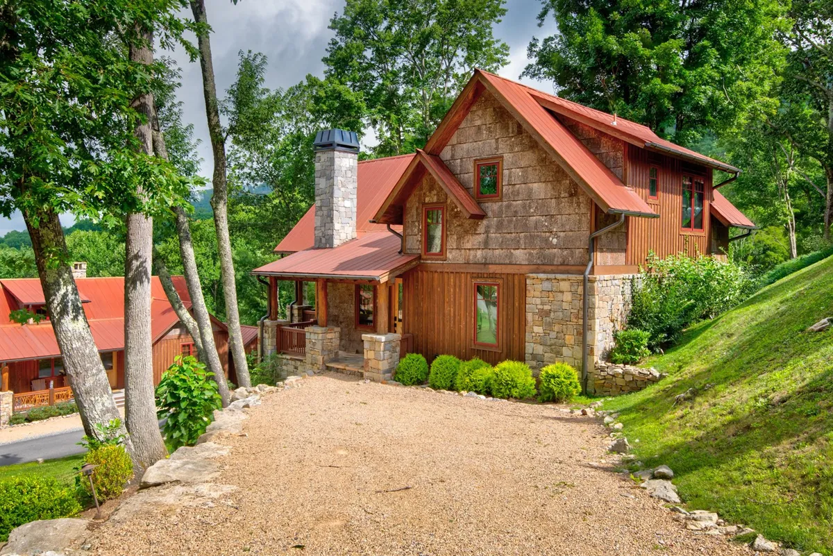 This exterior image features the cozy wooden facade of an Eagles Nest cottage, complete with a stone chimney, red metal roof, and surrounded by lush trees. The porch invites visitors to sit and enjoy the fresh mountain air, while the setting hints at the changing seasons with the vibrant greenery and natural landscape.